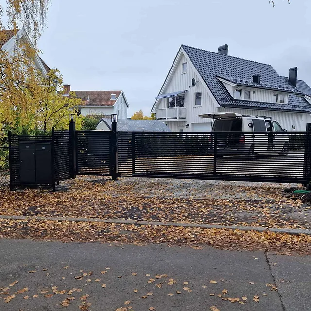 Stor, sort skyveport med lameller montert foran bolig med høstløv på bakken.
