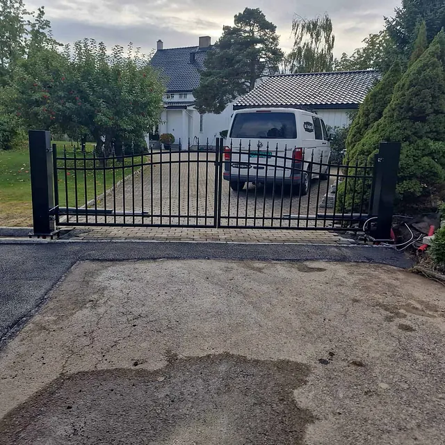 Innkjørsel med sort grind montert mellom steinsatte søyler foran hvitt hus med bil.